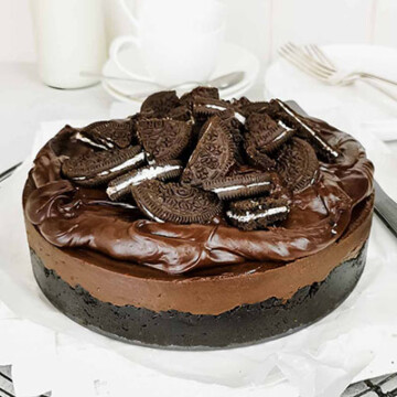 Chocolate cheesecake topped with chocolate frosting and chopped oreo cookies, against a white surface.