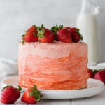 Strawberry cake on a white plate, topped with fresh strawberries, in front of a milk bottle.