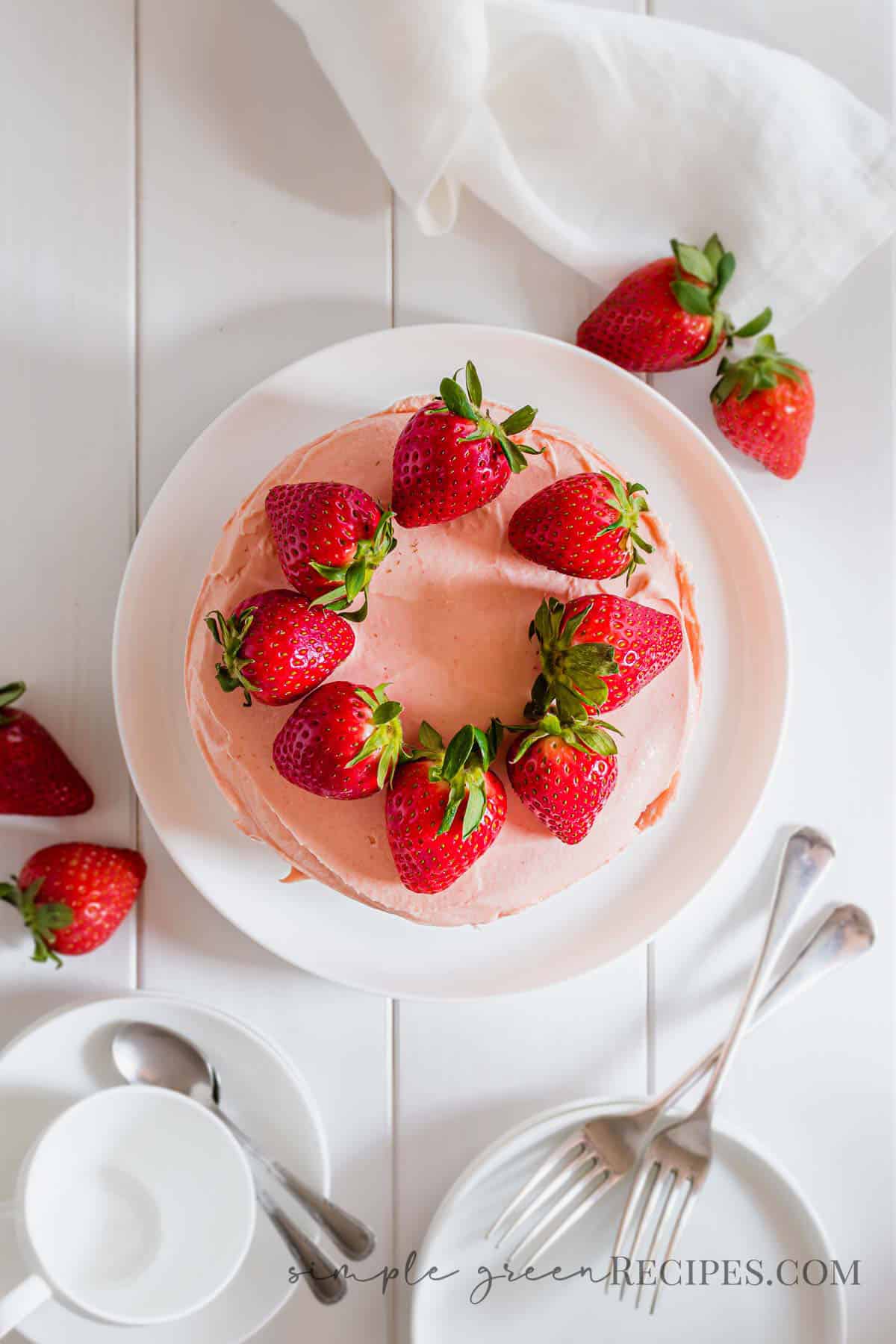 Strawberry cake on a white plate, topped with strawberries, next to dessert plates and coffee mugs.