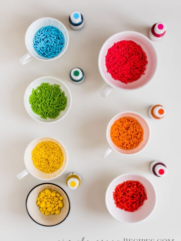 Seven white small bowls, with colorful homemade sprinkles, and the seven bottles of food coloring used.
