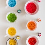 Seven white small bowls, with colorful homemade sprinkles, and the seven bottles of food coloring used.