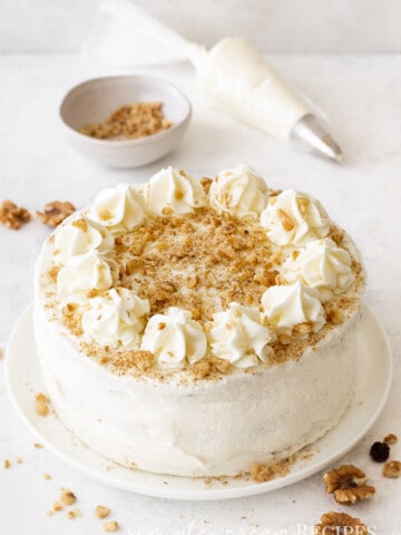 frosted carrot cake on a white plate, a bowl with walnuts and a piping bag